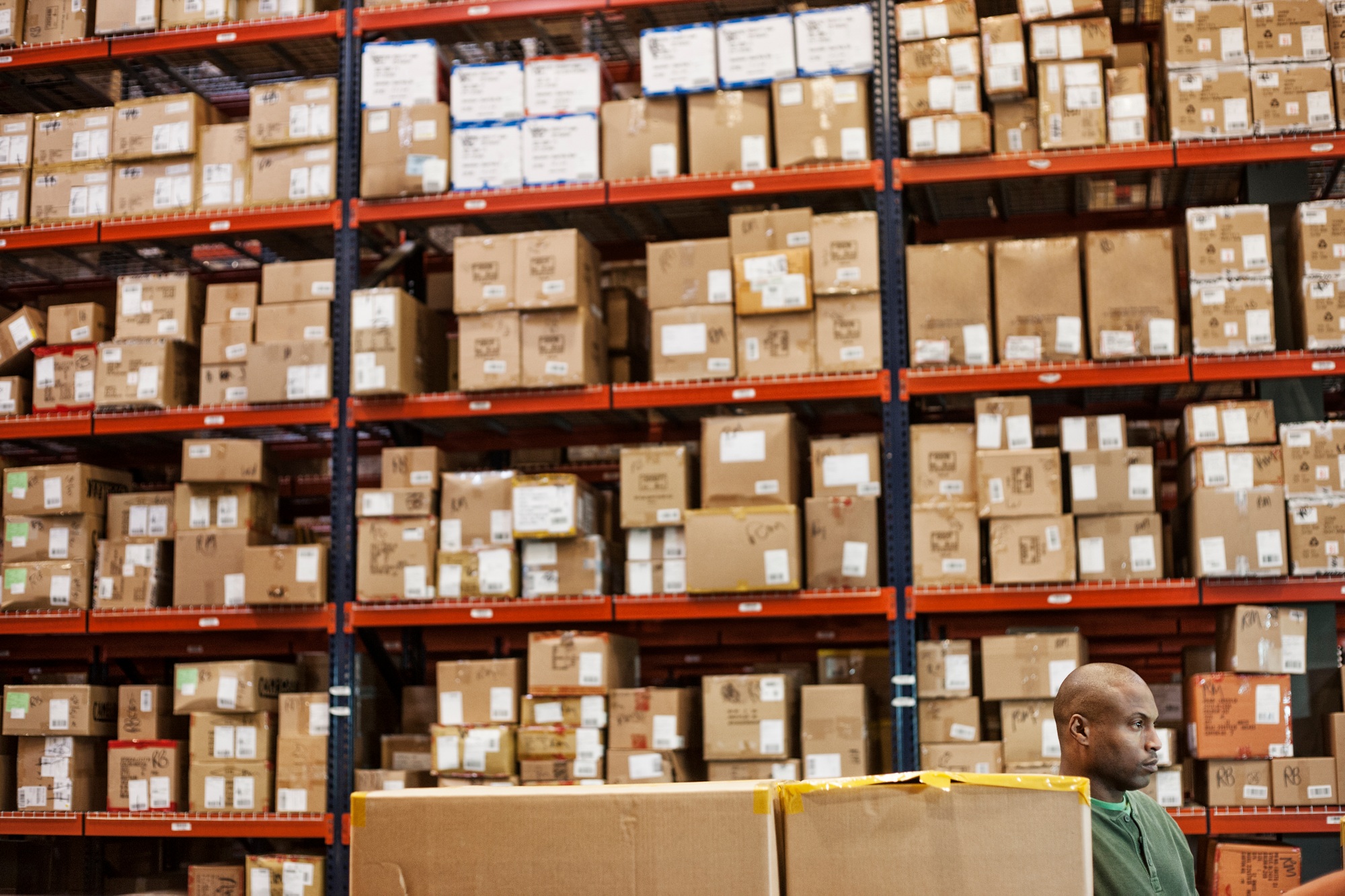 African American warehouse worker checking inventory next to large racks of cardboard boxes holding
