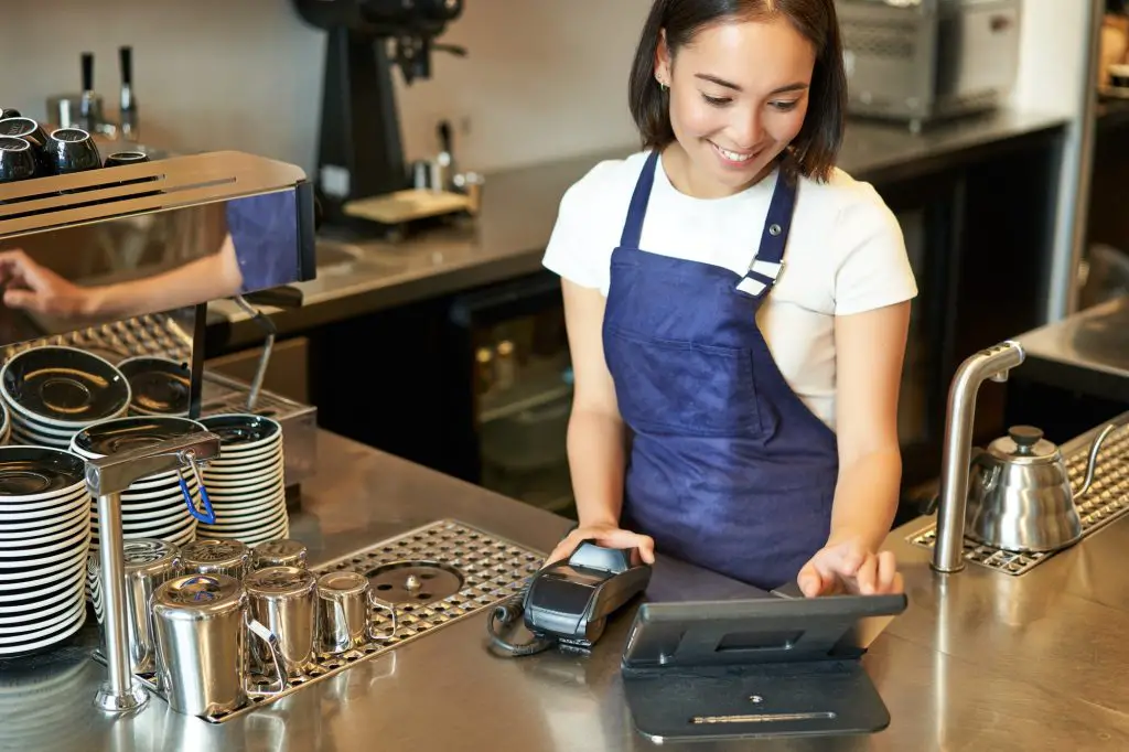 Smiling asian girl barista working in cafe at counter, processing orders, using POS terminal