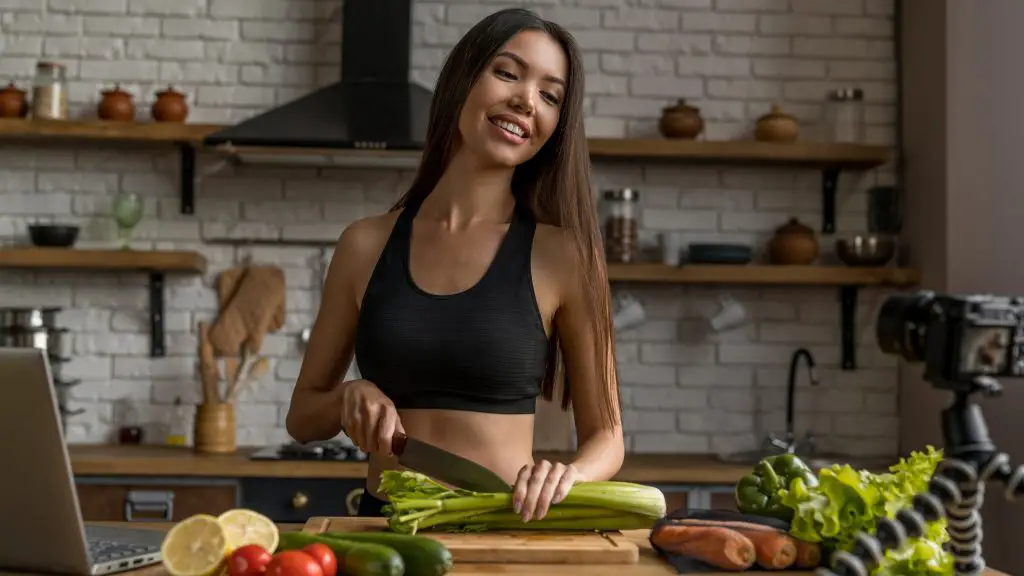 Smiling woman cutting celery in kitchen and recording it on video