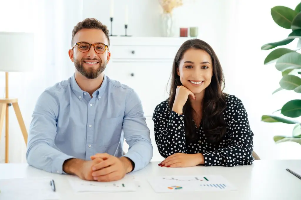 Successful confident man and woman sit side by side at a desk in the office, smiling at the camera