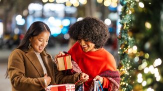 Two smiling young women exchanging christmas gifts in a decorated city street