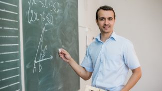 Young teacher or student holding triangle pointing at chalkboard with formula, looking to camera and